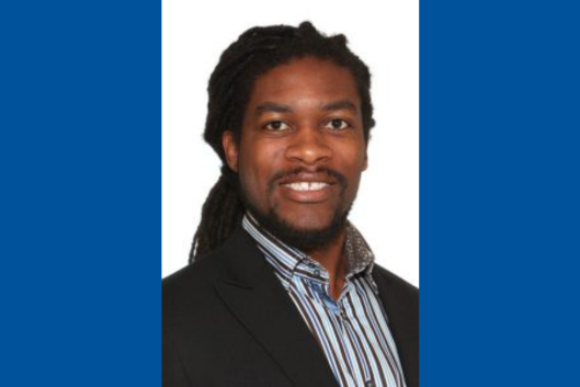 Pictured is Dr. Cavin Ward-Caviness, PhD, a smiling man with dark skin, long dark hair, and a well-groomed beard. He is wearing a black blazer over a striped dress shirt with a patterned collar. The background is plain white.